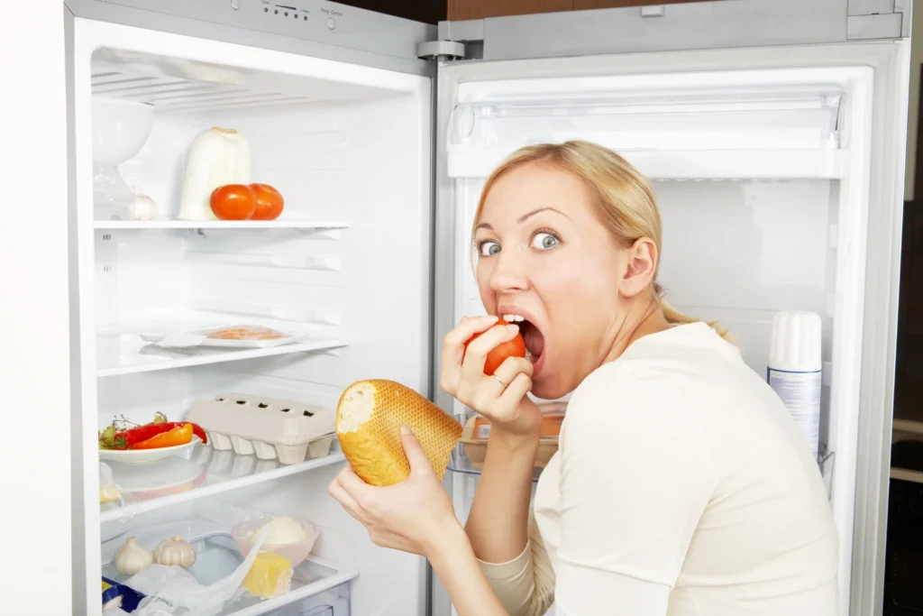 A woman is snacking from the fridge; she can't control her hunger.