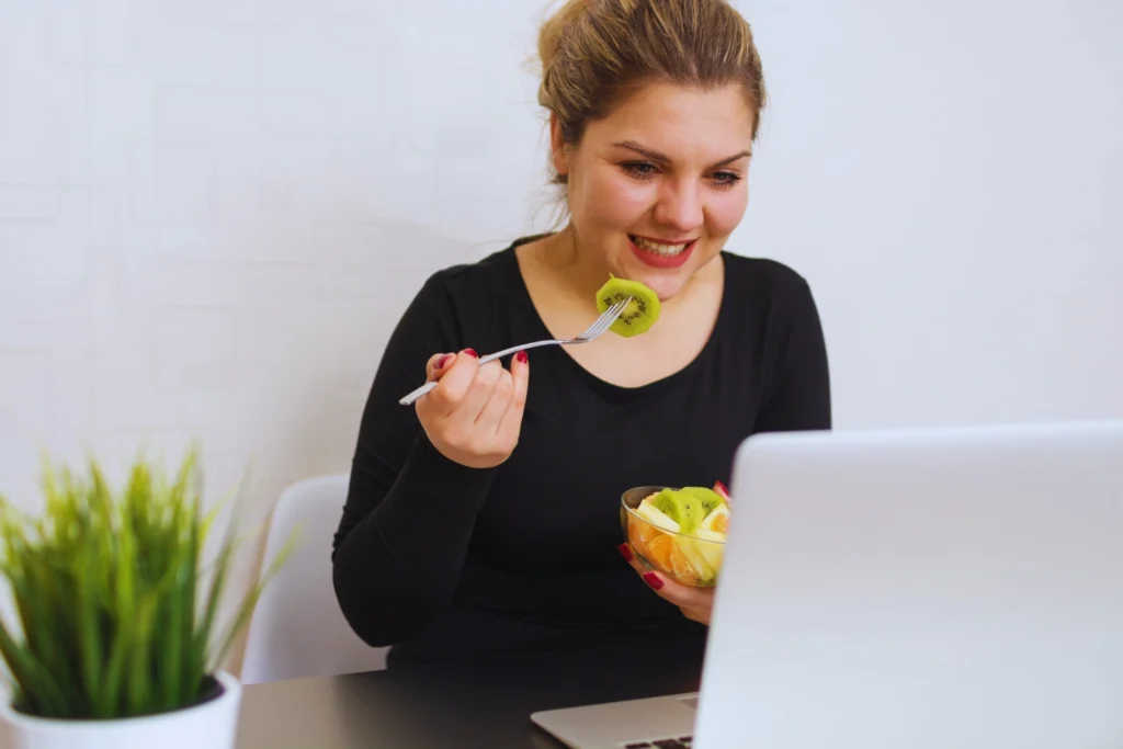 A woman is eating a fruit salad and reading research on metabolism.
